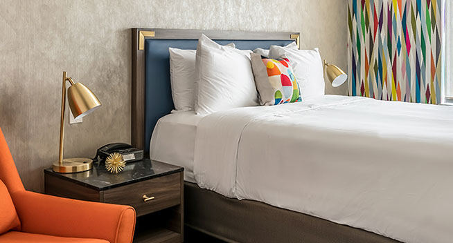 Queen Guest Room with Sleeper chair. This photo shows the colorful ambience with a queen size bed and an orange sleeper chair, nightstand and lamp with alarm clock.