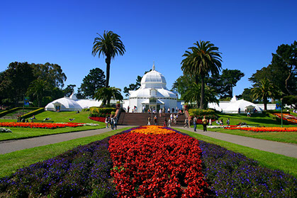 view of Conservatory of Flowers in the Golden Gate Park. Beautiful red and orange flowers mixed with purple line the grass ways.
